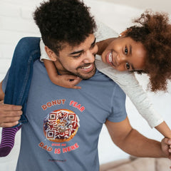 Dad in a blue t-shirt with a donut-themed QR code design reading “Donut Fear, Dad Is Here,” giving his daughter a piggyback ride indoors.